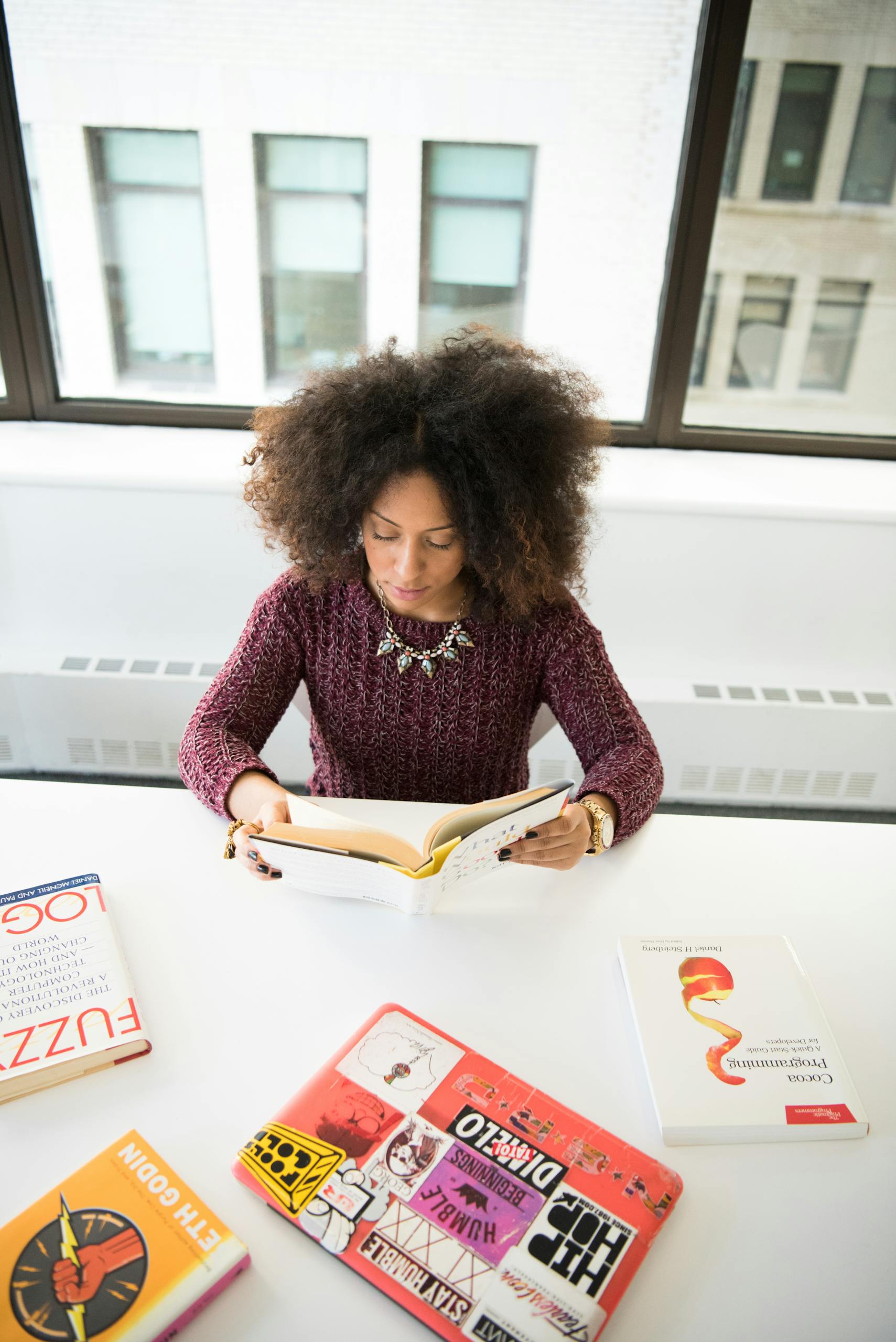 A woman engaged in reading a book at a white desk, surrounded by colorful books, in a modern office space.